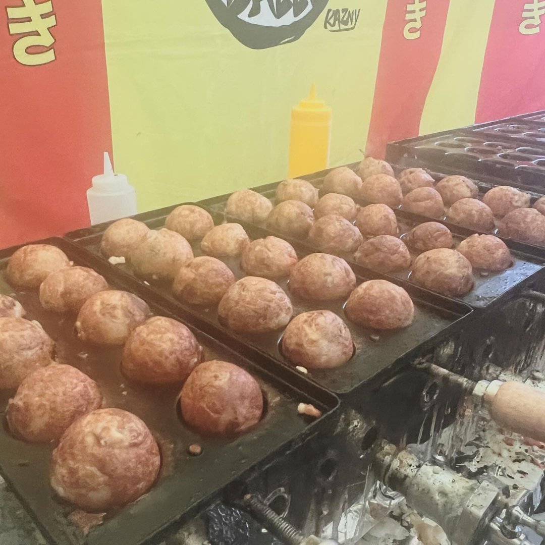 Rows of takoyaki cooking on griddle plates at a KARLSBALLS event, the shop banner behind